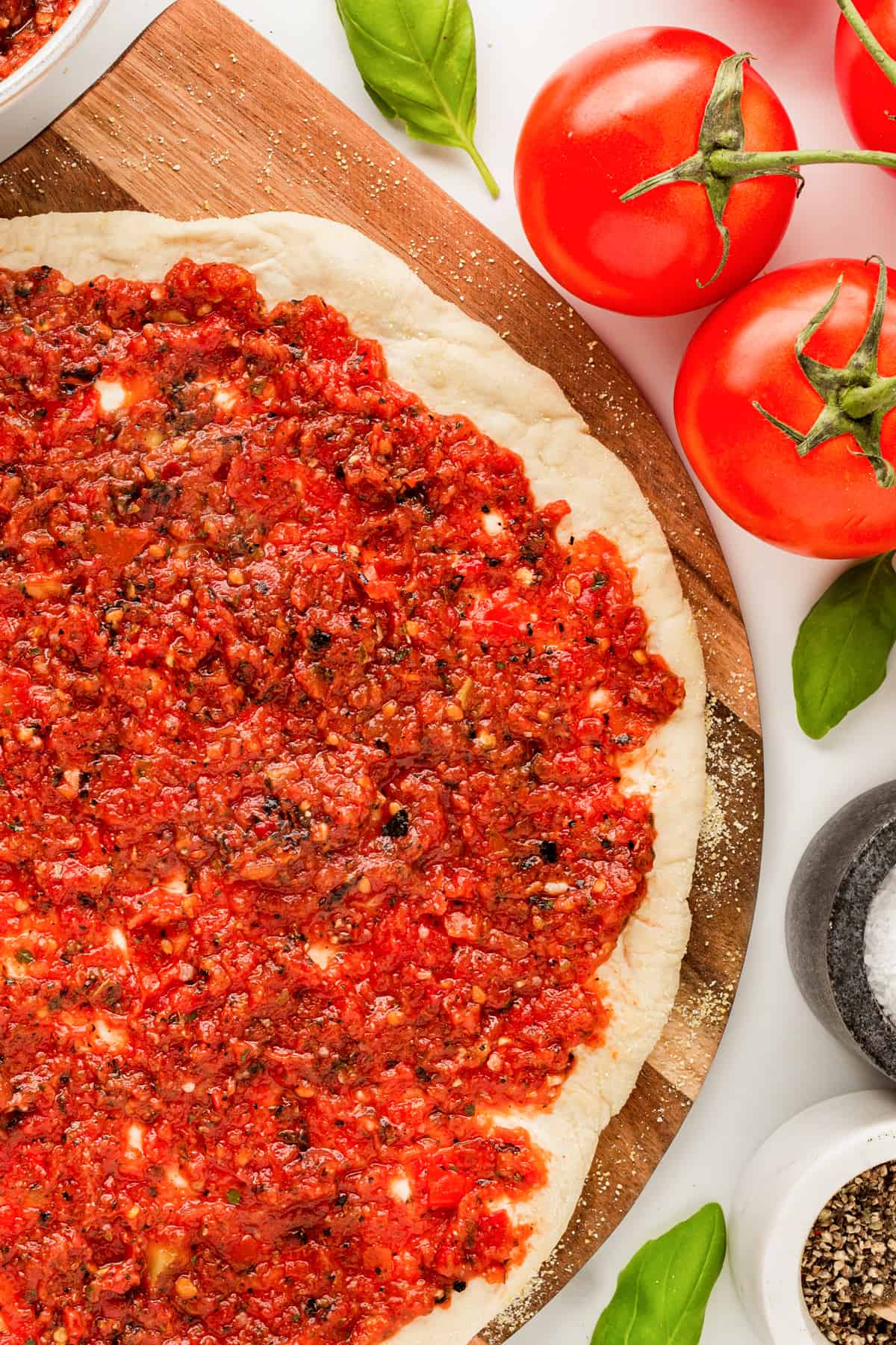 Homemade pizza sauce on a pizza dough with fresh tomatoes and basil on the side. 