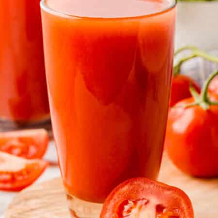 Glass of tomato juice with half a slice of tomato in front.