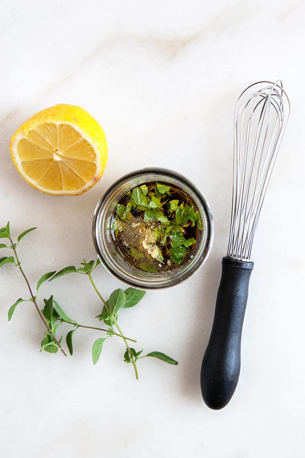 A whisk beside a small jar of dressing with a lemon and oregano. 