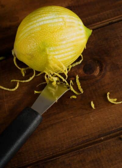 Partially zested lemon with a zester beside it on a dark wood background.