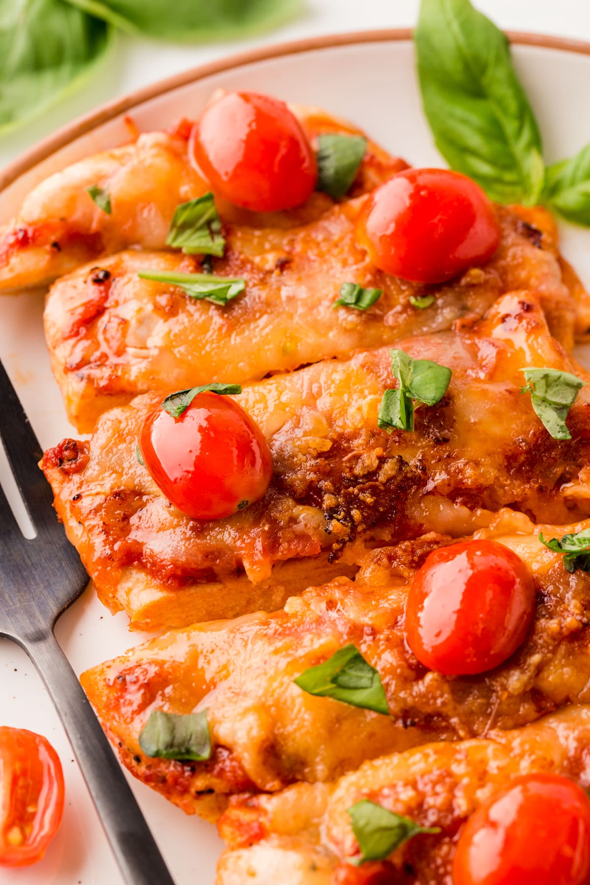 Close-up picture of sliced pizza chicken on a plate, topped with sliced cherry tomatoes and basil. 