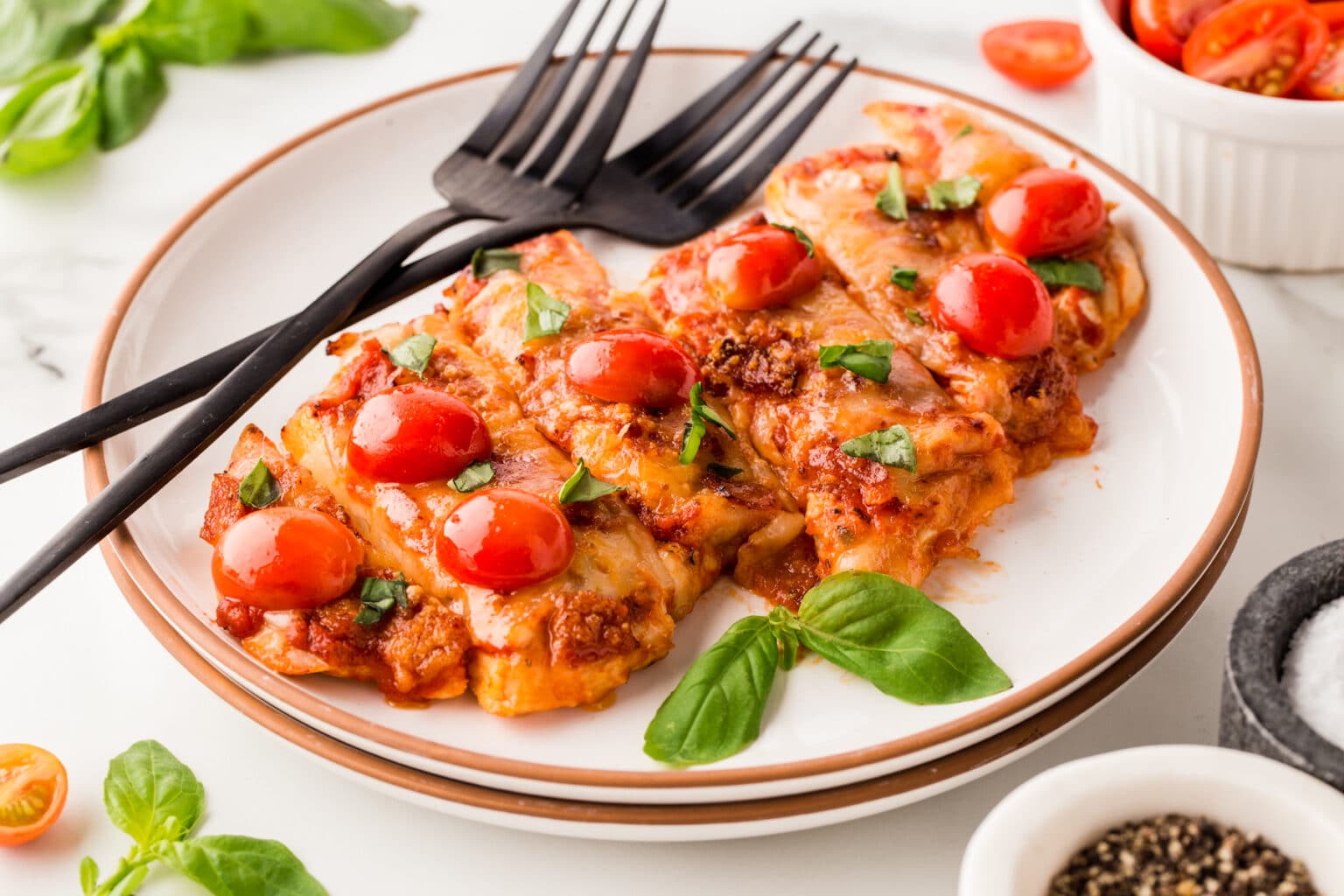 A plate of lasagna topped with cherry tomatoes and fresh basil leaves. Two black forks rest on the plate. In the background, there is a small bowl of cherry tomatoes and a mortar containing salt and pepper. Fresh basil leaves surround the scene.