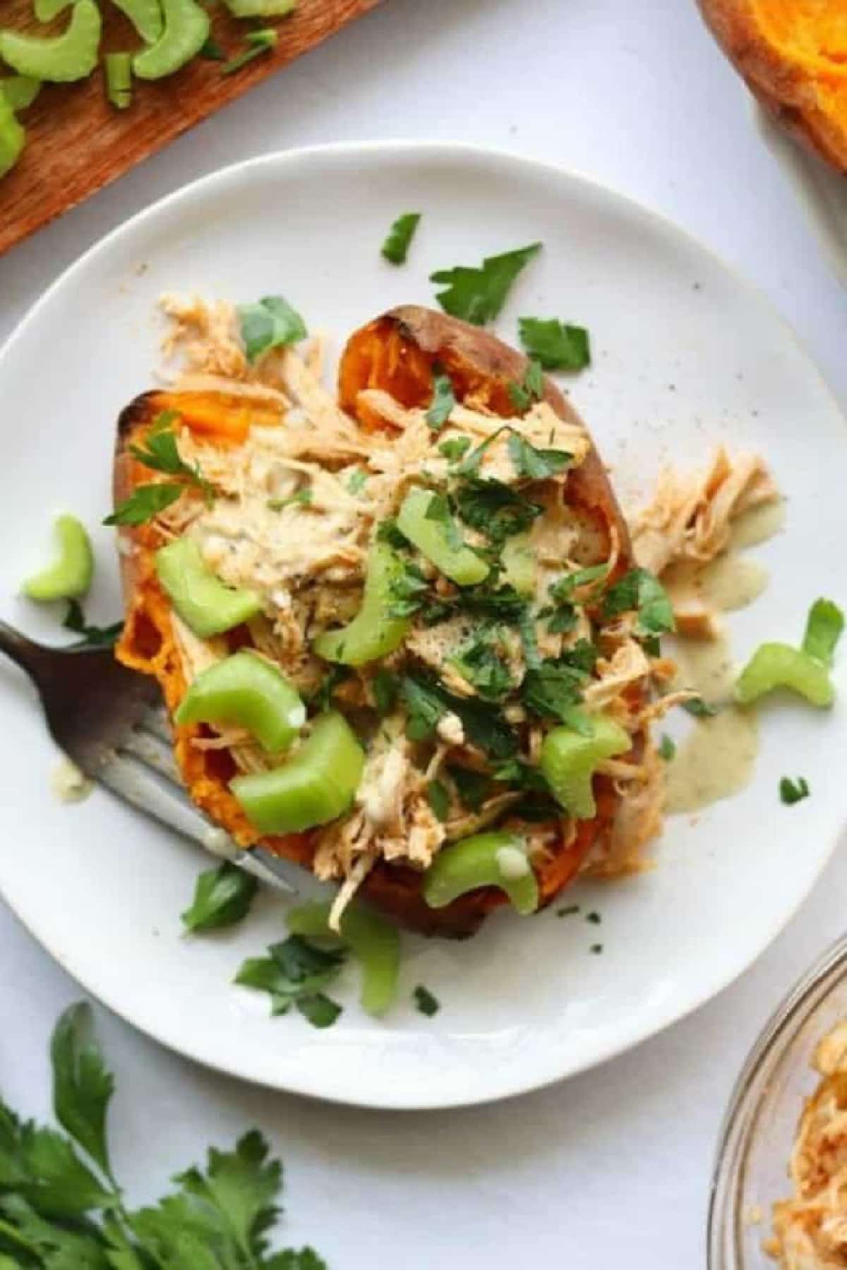 Instant Pot Buffalo Chicken Stuffed Sweet Potato on a white plate with a fork and sliced celery and parsley for garnish.
