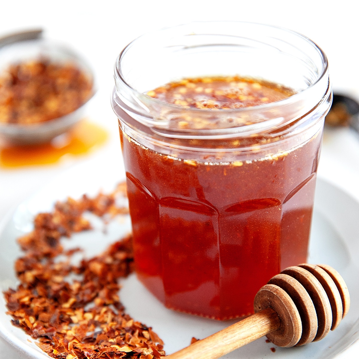 Square photo of hot honey in a small jar with a honey dipper and lots of chili flakes around the jar. 