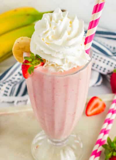 Strawberry Banana Milkshake in a glass with whipped cream, straw and sliced fruit on the side.