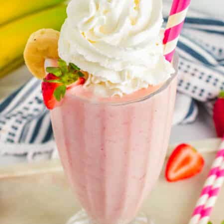 Strawberry Banana Milkshake in a glass with whipped cream, straw and sliced fruit on the side.