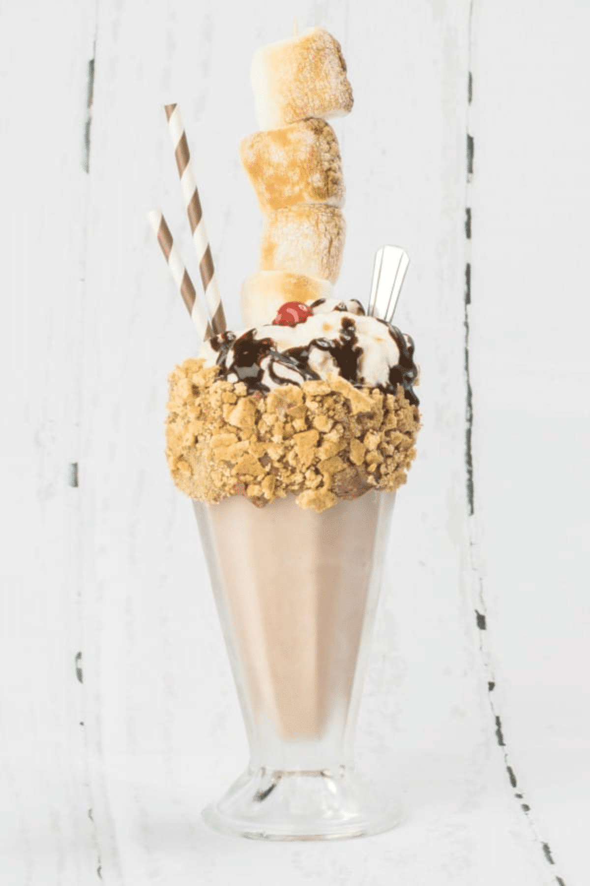 S'mores milkshake in a milkshake glass with a graham cracker rim, chocolate sauce and a cherry on top with browned marshmallows on a stick and straws and a spoon. 