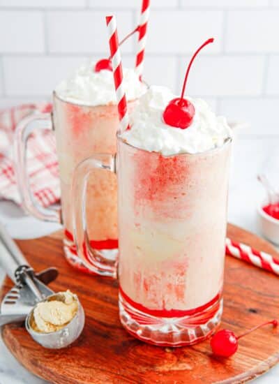 Two cherry floats with straws on a board with an ice cream scoop beside them.