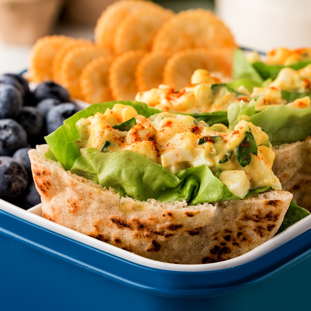 Square photo of egg salad pitas in a lunchbox showing the first one close-up. 