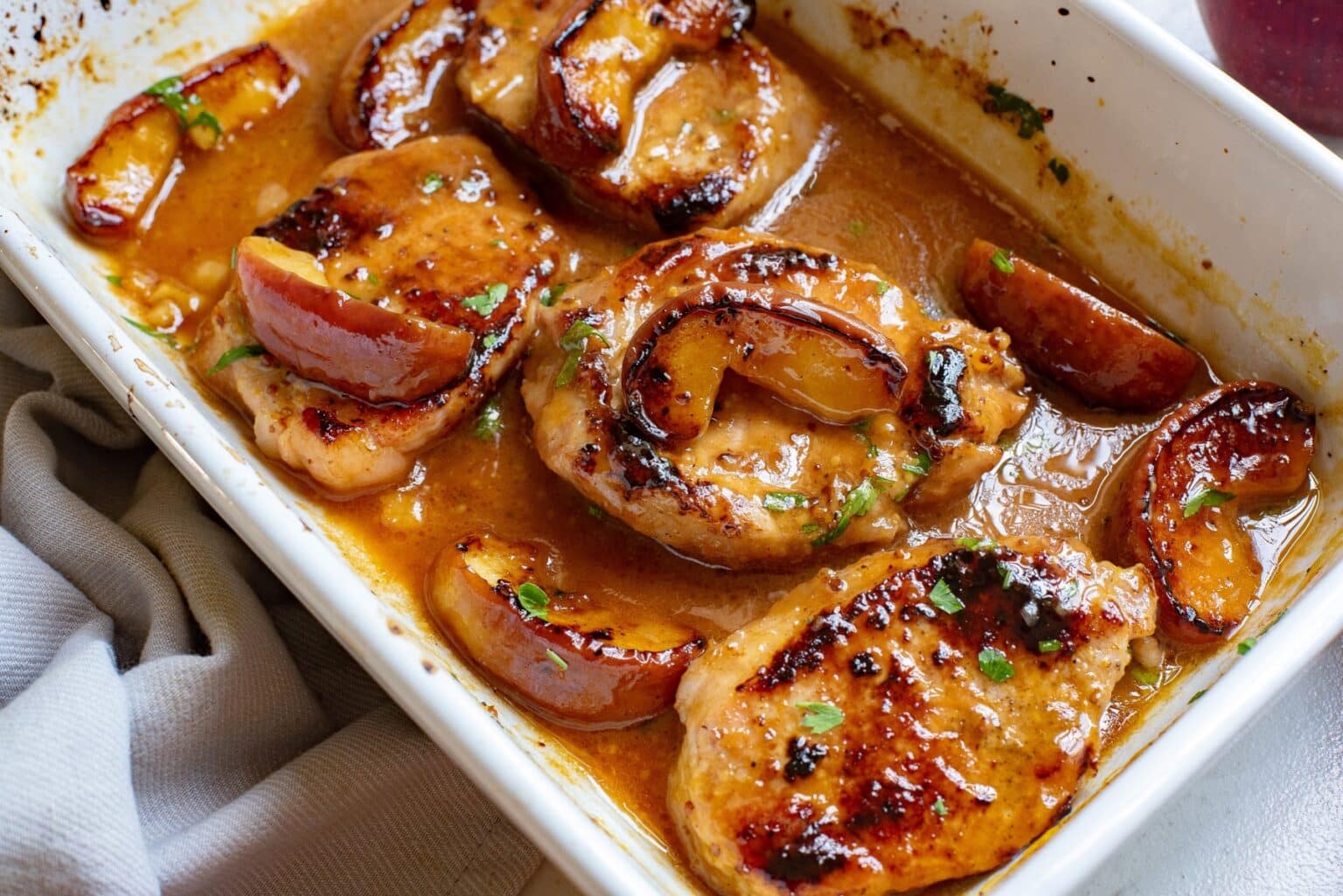 A close-up of a white baking dish filled with glazed pork chops and caramelized apple slices in a rich, golden-brown sauce. The pork chops are garnished with small green herbs and look juicy and well-cooked. The dish is placed next to a light beige cloth napkin.
