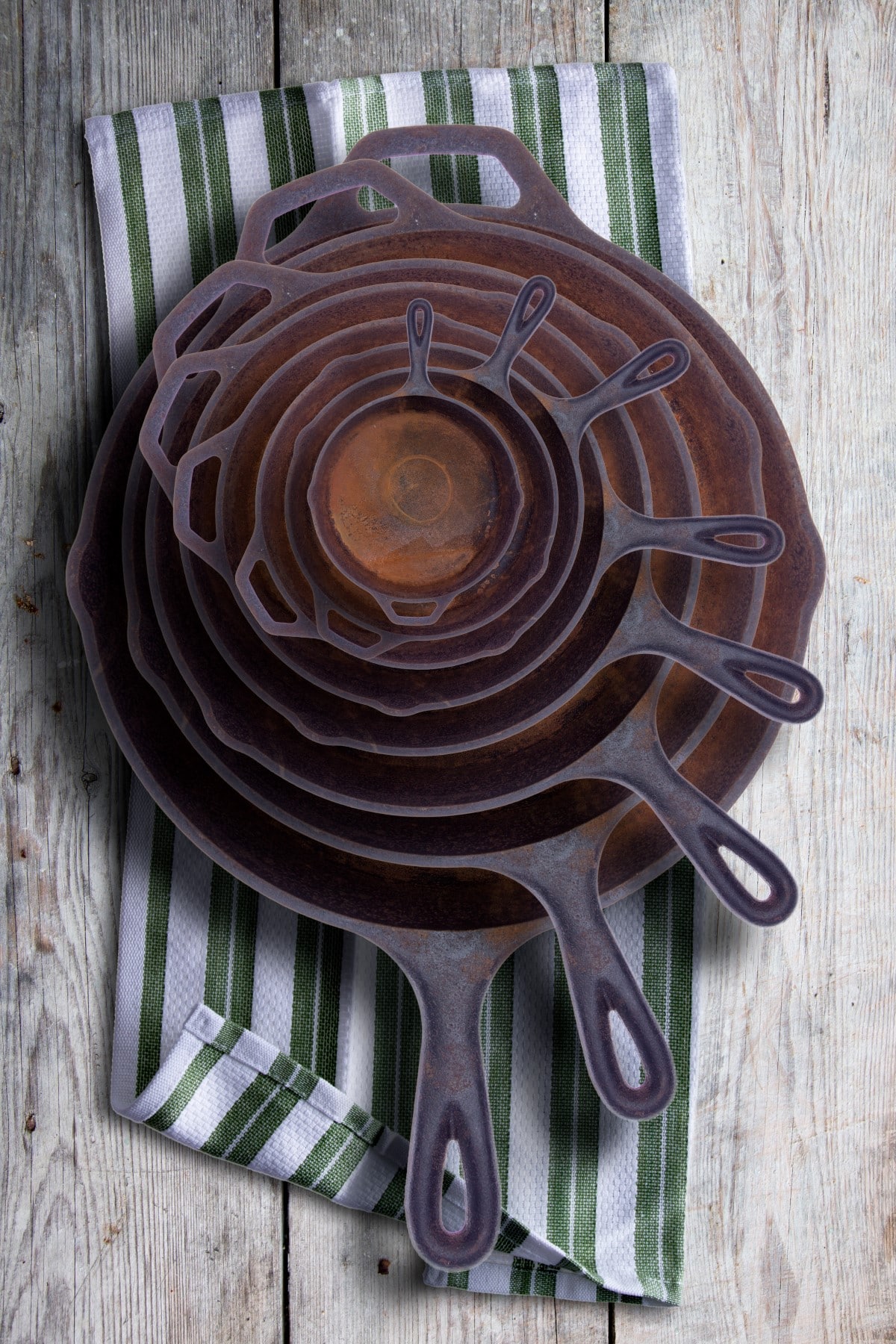 Collection of round rusty cast iron frying pans in diminishing sizes stacked one inside the other, viewed from above on a rustic napkin on an old wooden table.