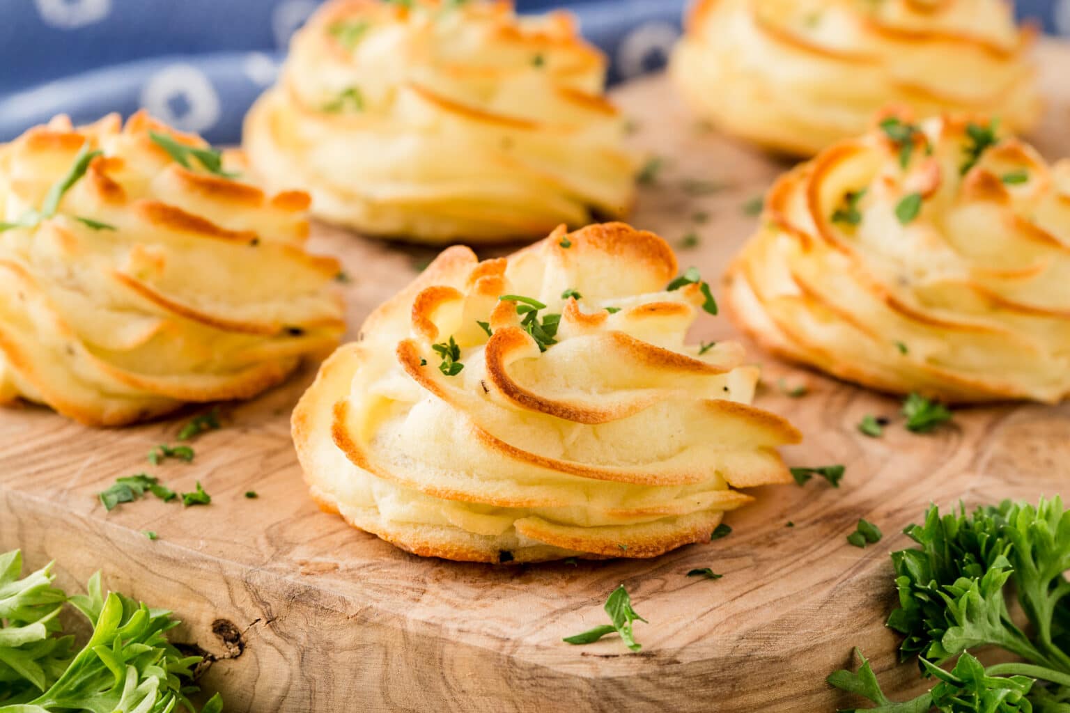 Golden brown duchess potatoes garnished with fresh parsley, arranged on a wooden board. These piped, baked potato mounds are set against a blue-patterned background, with sprigs of parsley around them.
