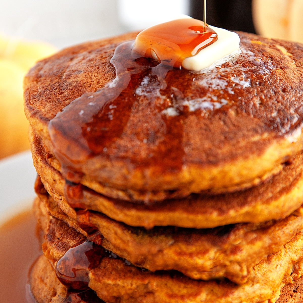 Square photo of a stack of pancakes with butter and maple syrup being poured on. 