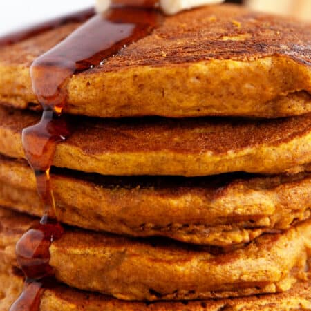 Stacked Pumpkin Pancakes with butter on top and maple syrup pouring on.