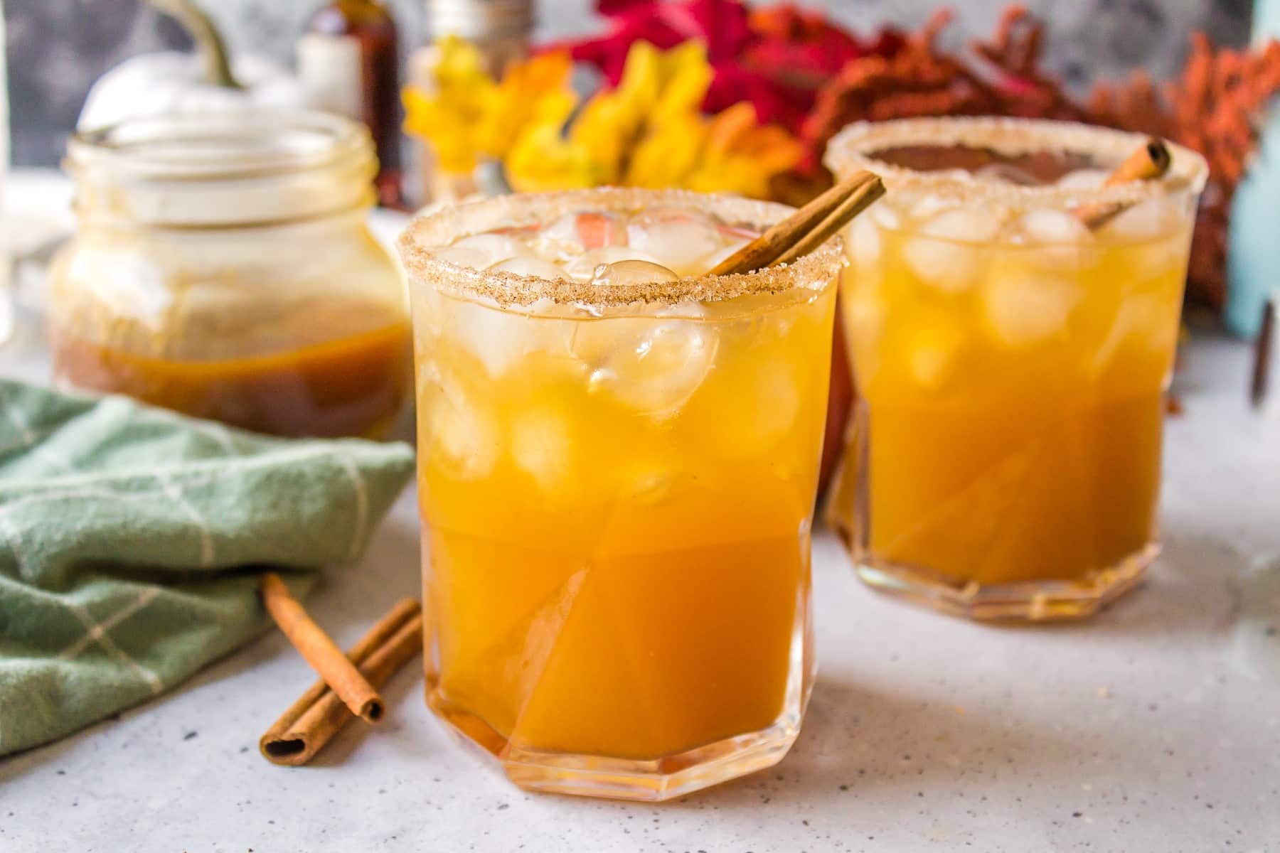 Horizontal picture of two cocktails with pumpkin puree and leaves and a light green kitchen towel. 