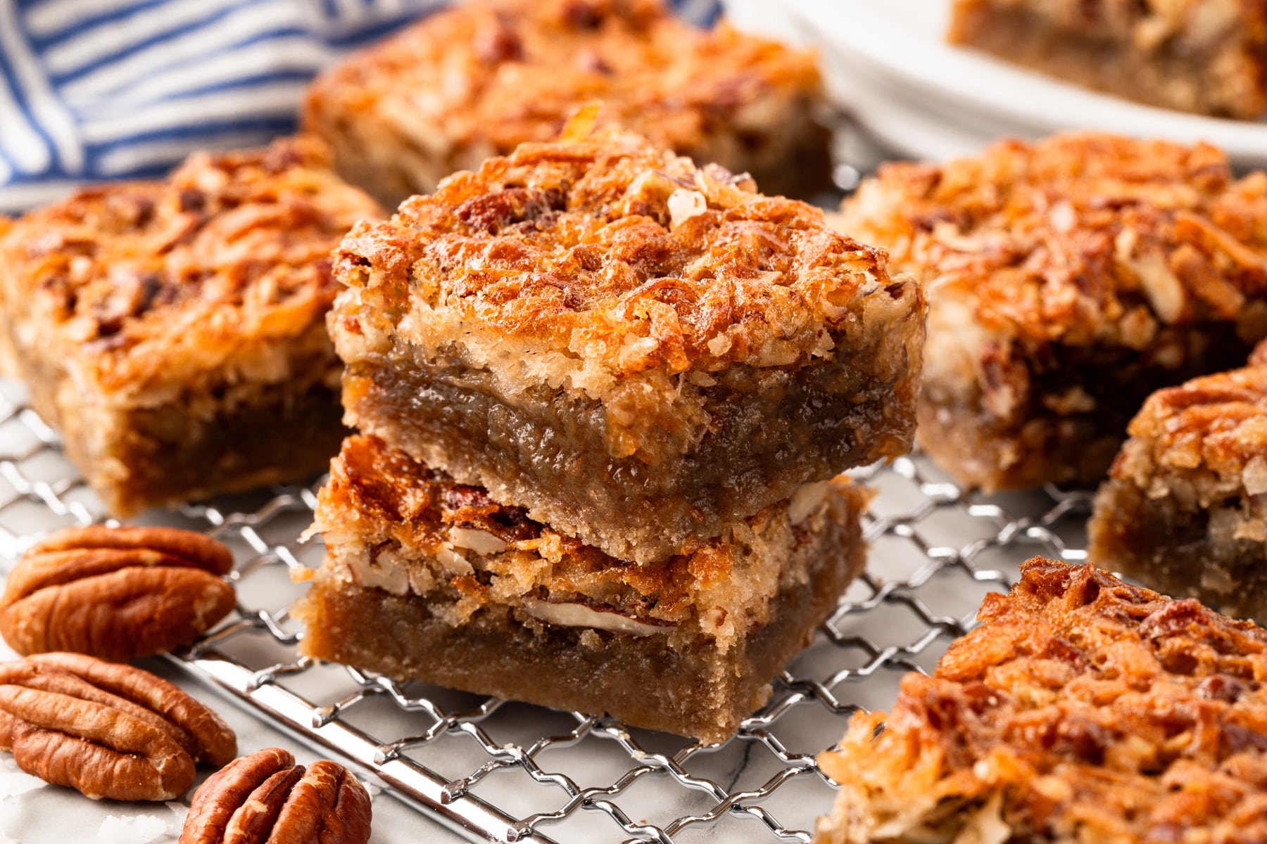 Horizontal picture of two pecan bars stacked and more around them on a cooling rack. 