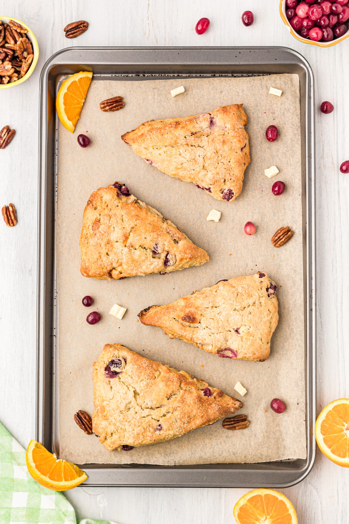 Scones on a parchment-lined baking sheet with cranberries, pecans and white chocolate chunks scattered. 