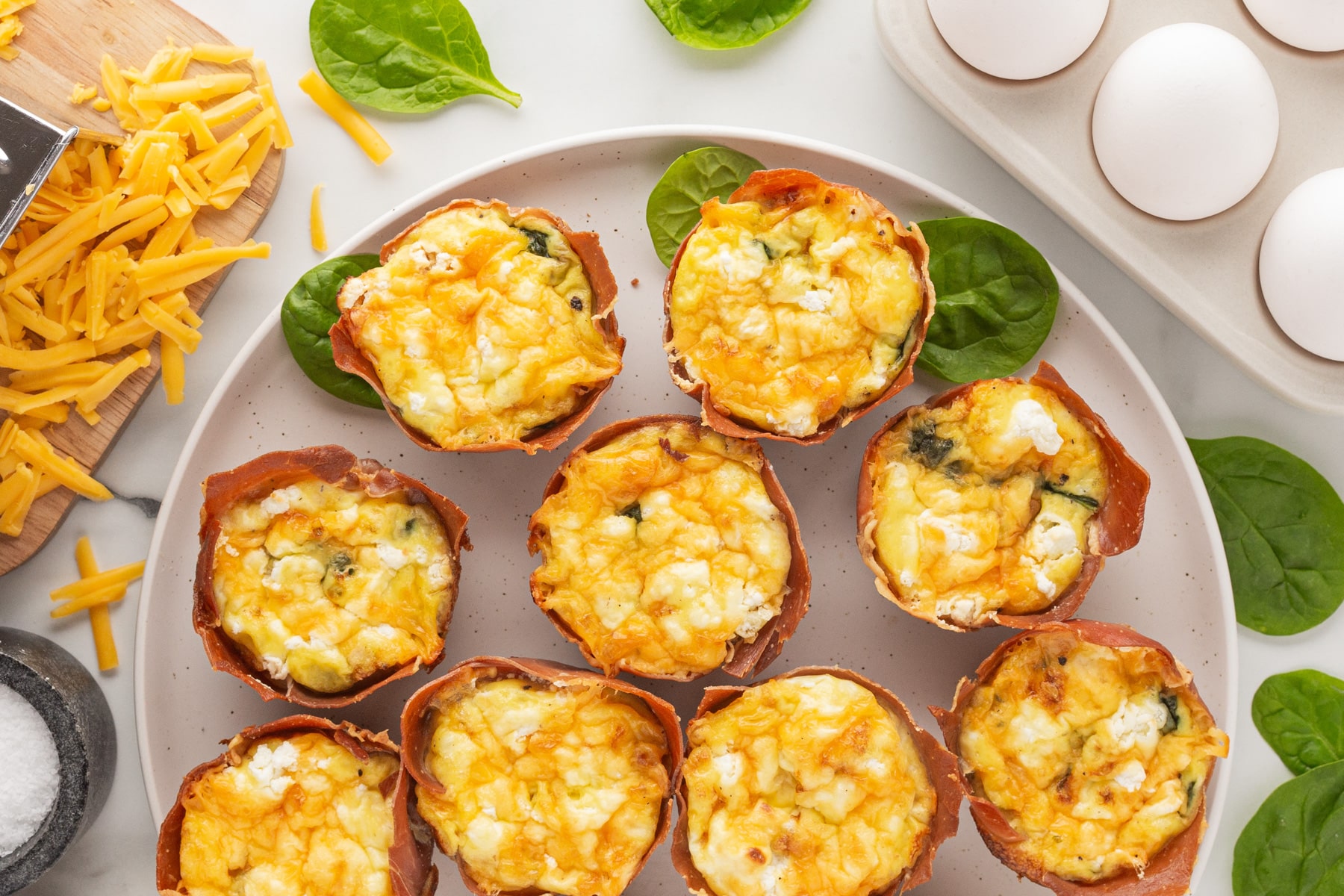 Horizontal pic of egg cups on a plate with whole eggs and grated cheese on the sides. 