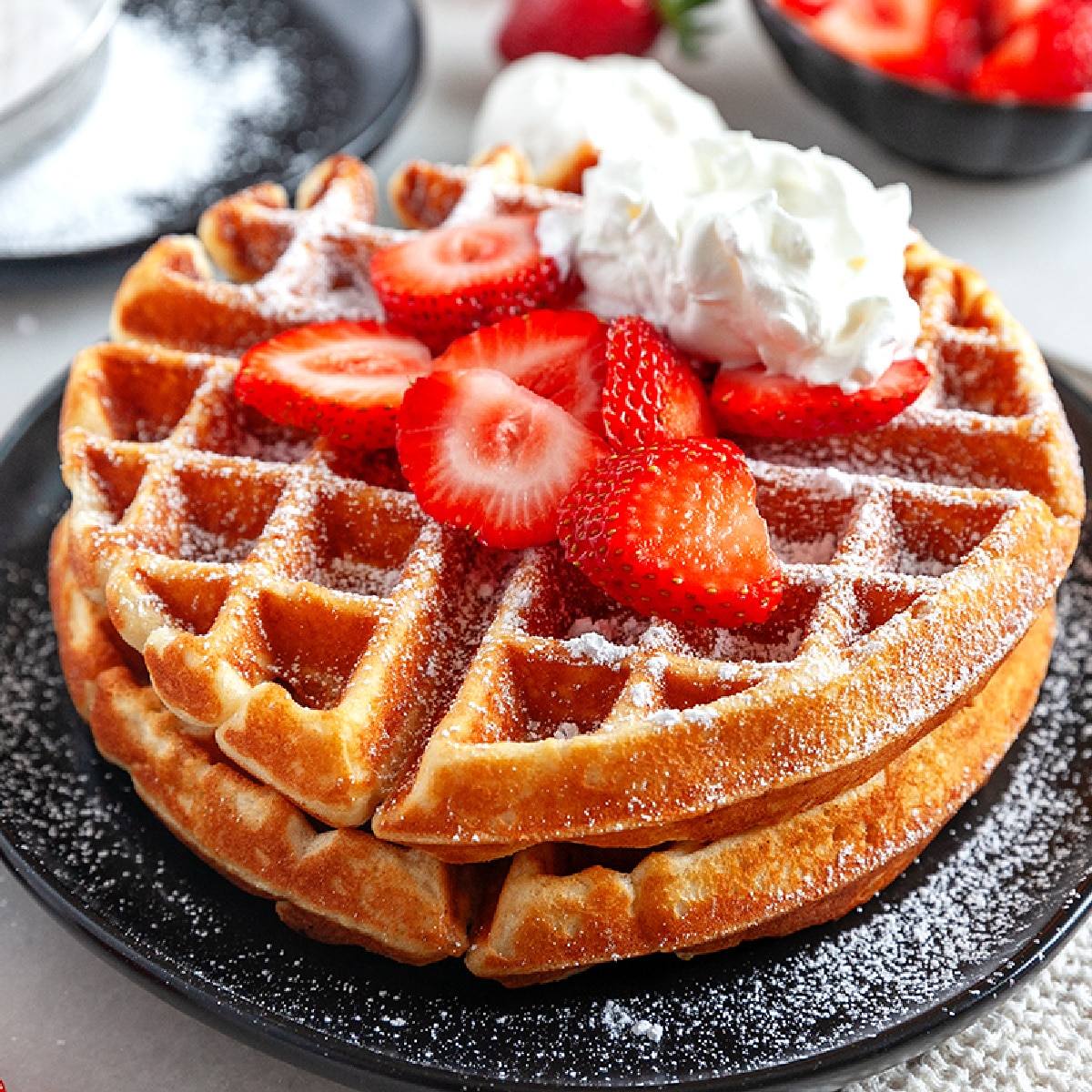 Square photo of Belgian Waffles topped with strawberries and whipped cream. 