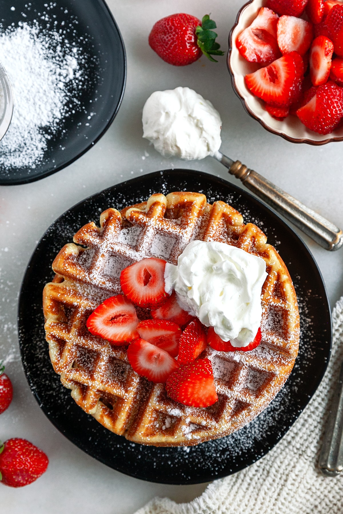 Overhead shot of a Belgian Waffle on a plate topped with strawberries, icing sugar and whipped cream. 