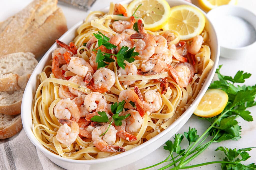 A white oval dish holds a serving of fettuccine pasta topped with shrimp, fresh parsley, and lemon slices. Nearby, there is a piece of crusty bread, several fresh lemon slices, and parsley sprigs. The dish is set on a white surface.