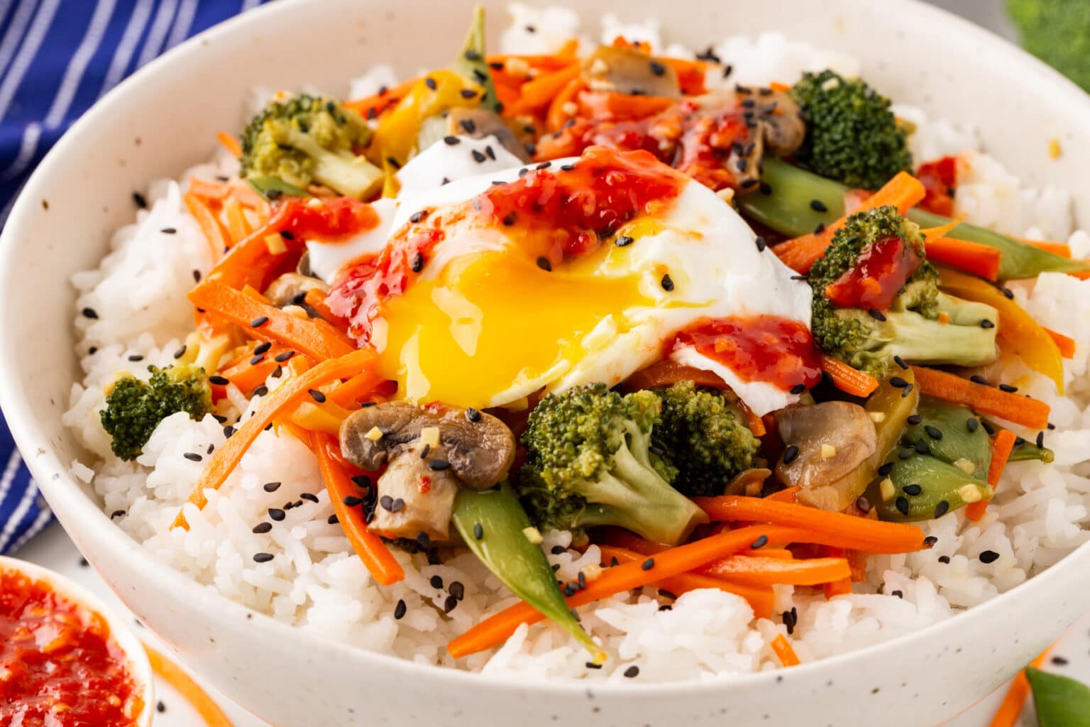 A bowl of rice topped with a runny poached egg, broccoli, carrots, mushrooms, and black sesame seeds. Drizzled with a red sauce, it sits in a speckled bowl on a table with a blue-striped napkin beside it.