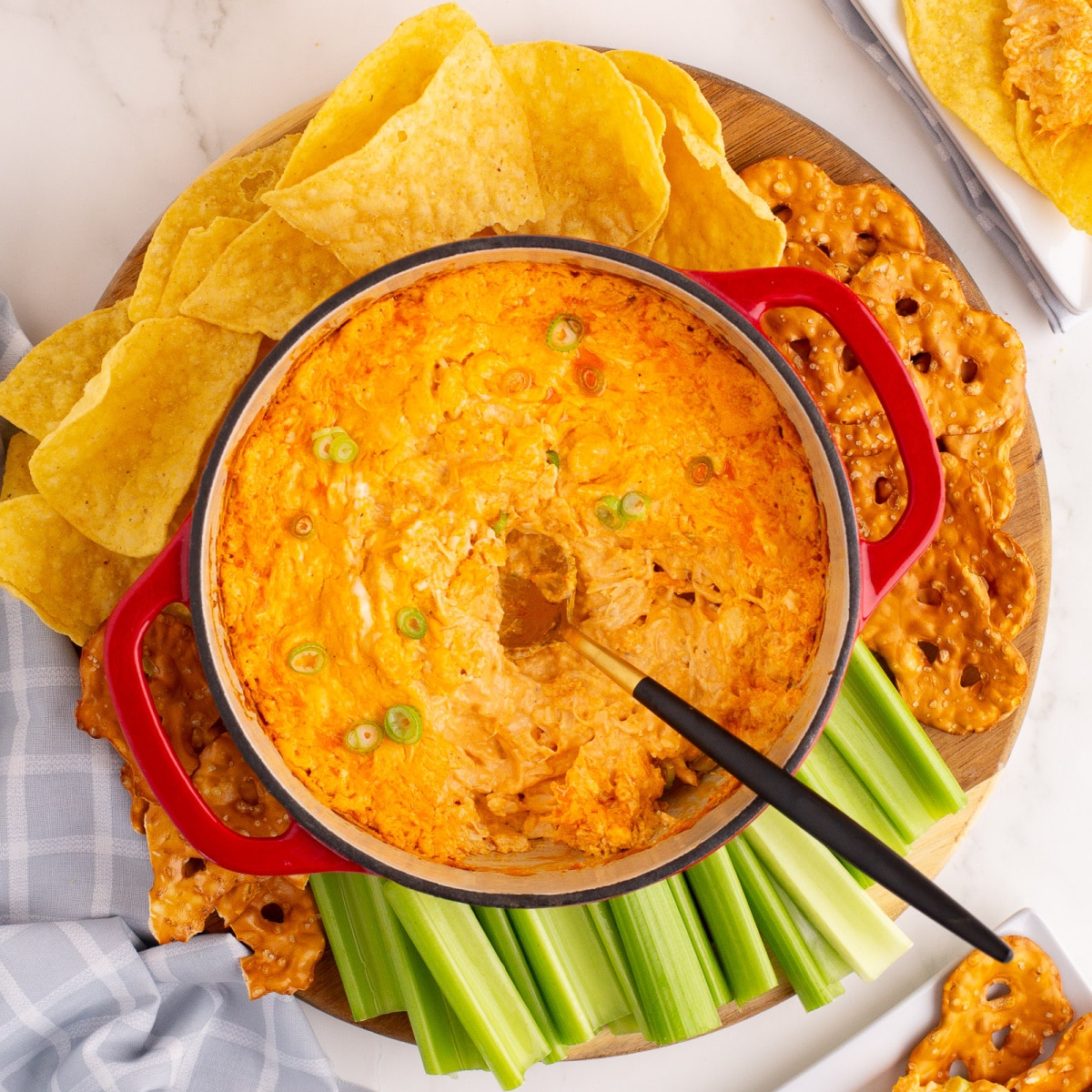 Square overhead shot of the Buffalo Chicken Dip in a round red casserole with a spoon in it and dippers like tortilla chips, celery and pretzel chips surrounding. 