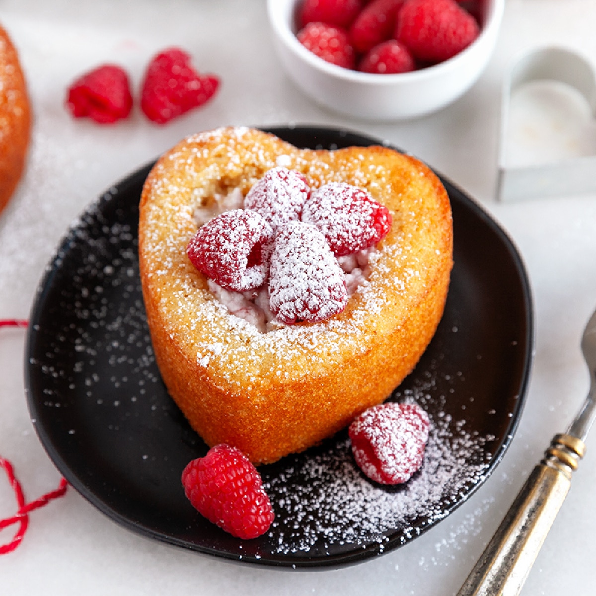 Heart shaped cake on a black plate with raspberries and dusted with icing sugar. 