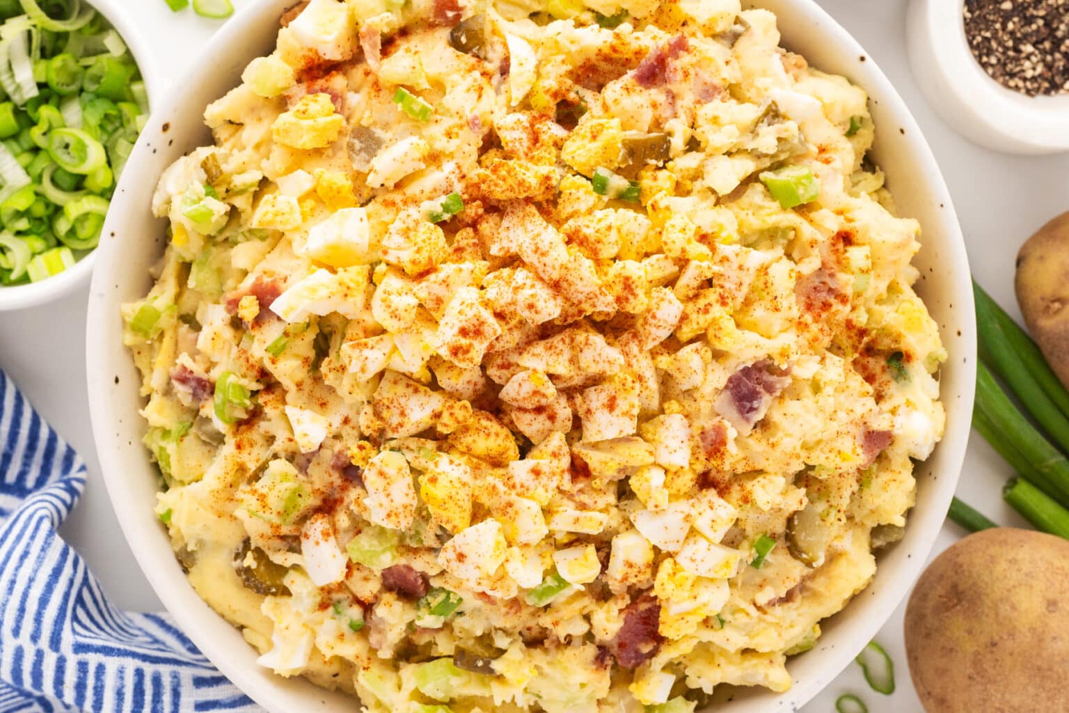 A bowl filled with creamy potato salad topped with chopped green onions and sprinkled with paprika. Surrounding the bowl are whole potatoes, green onions, and a white and blue striped cloth. Other small dishes are partially visible in the background.