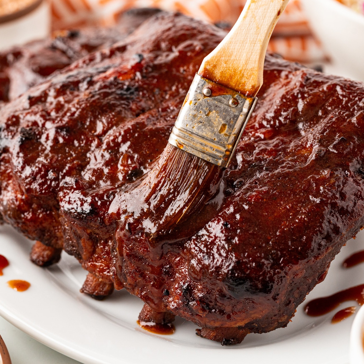 Square photo of ribs being brushed with BBQ sauce. 