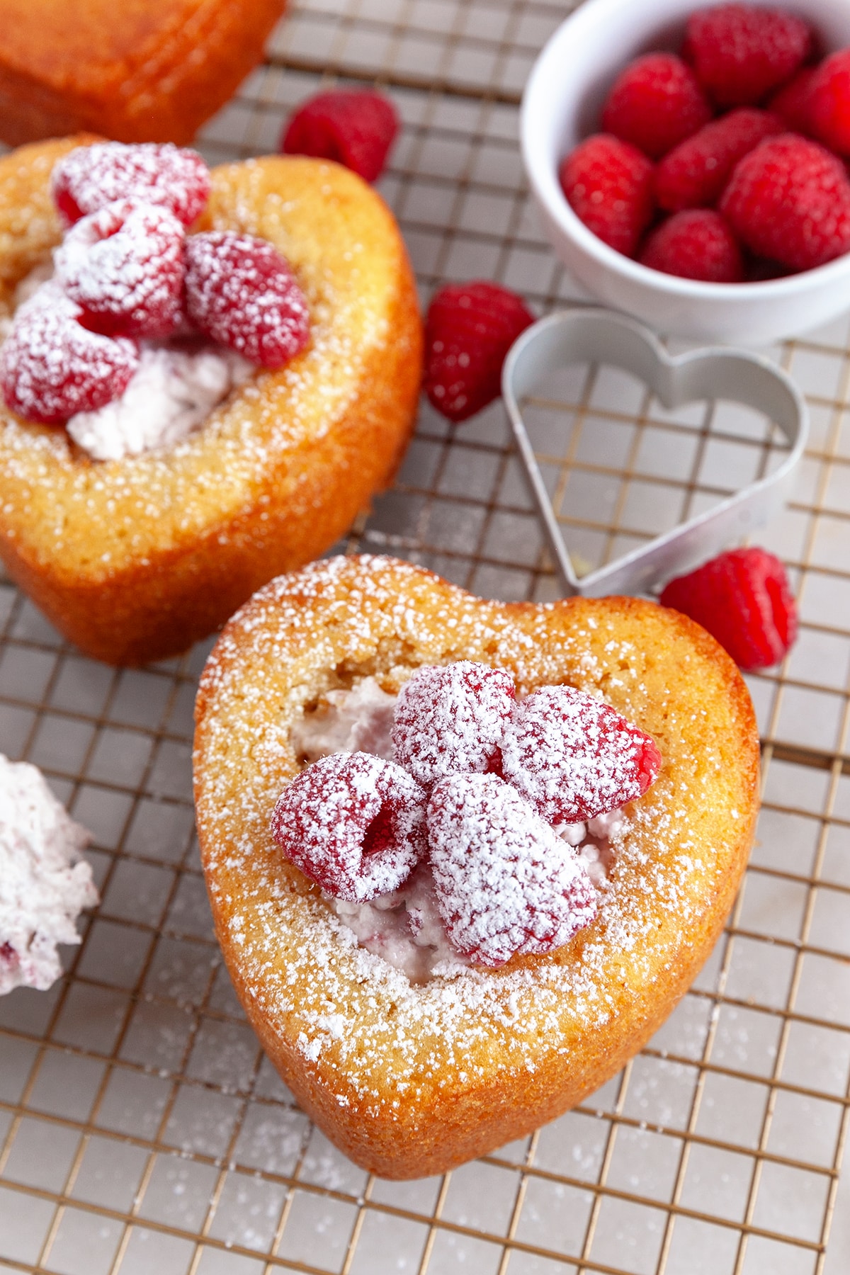 A heart shaped cake on a cooling rack topped with raspberries and dusted with icing sugar. 