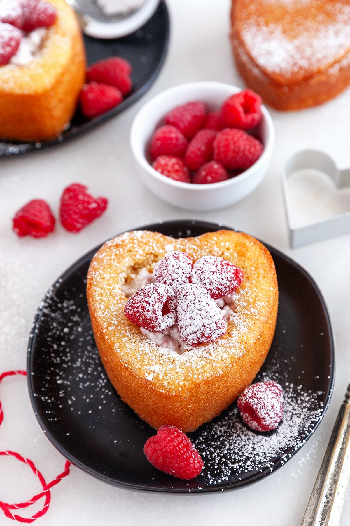 Heart shaped cake on a black plate with raspberries and dusted with icing sugar. 