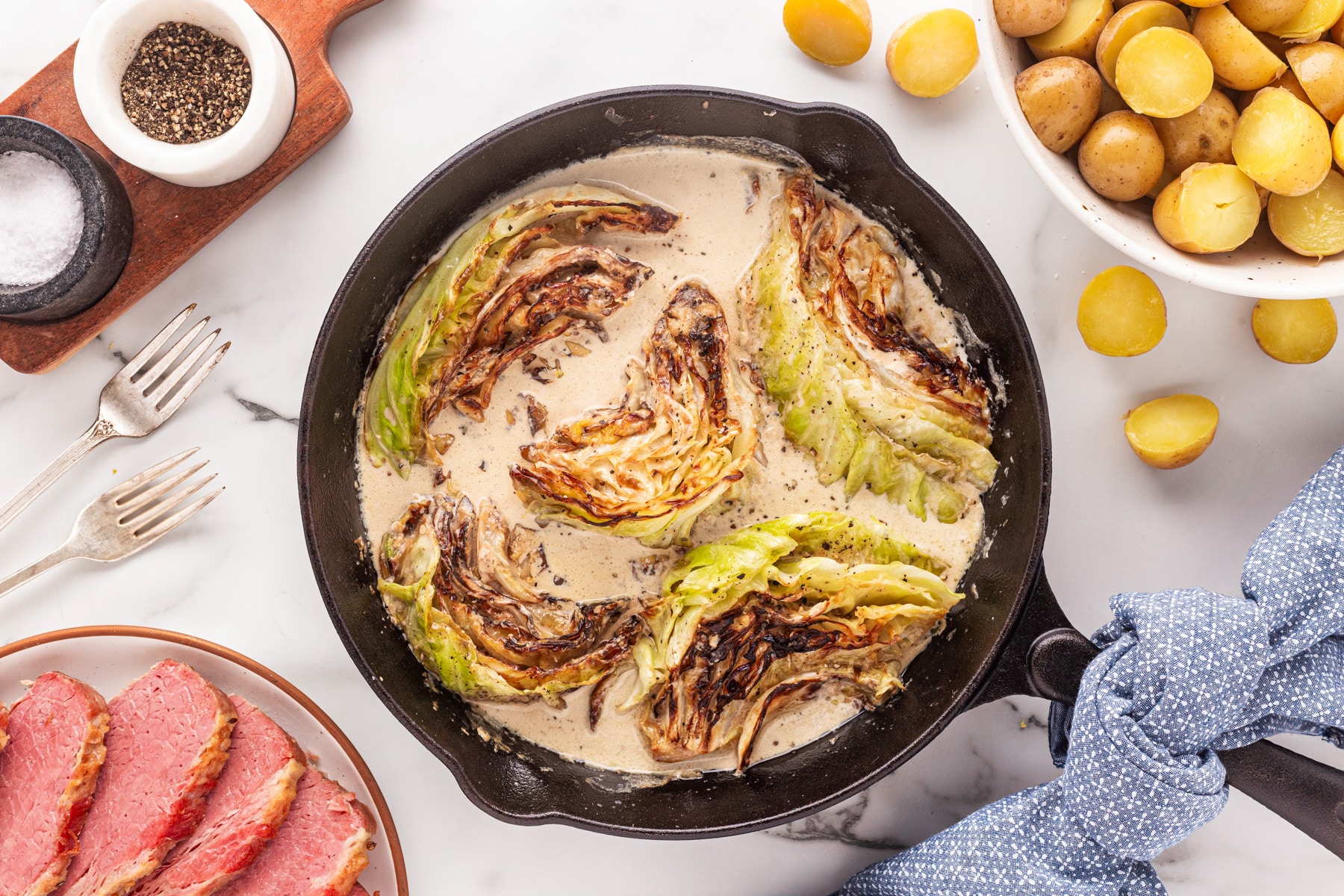 Horizontal picture of creamed cabbage in a cast iron pan with corned beef and potatoes on either side at angles. 