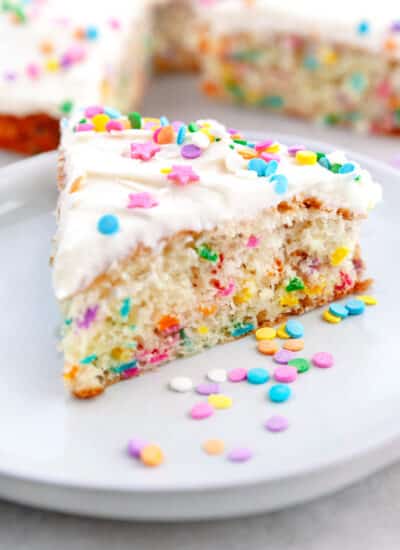 Showing a slice of Funfetti Cake on a white plate with confetti on the plate.
