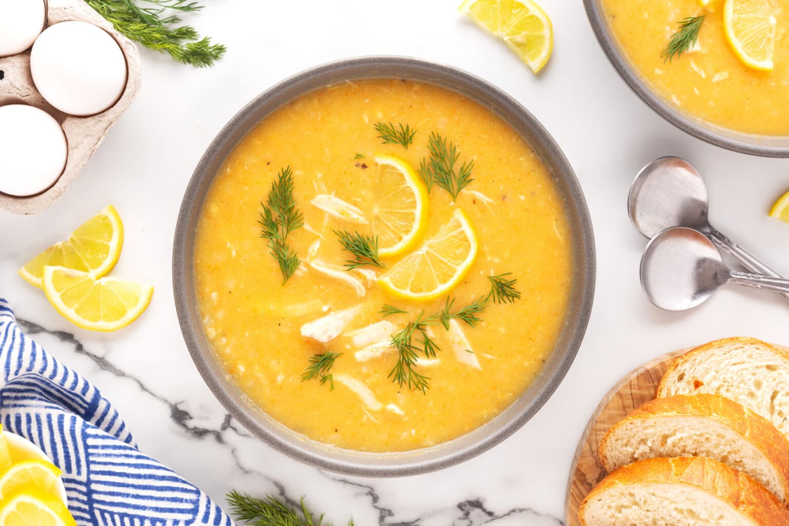 A bowl of creamy yellow soup garnished with lemon slices and fresh dill, accompanied by slices of bread, two spoons, and lemon wedges. Near the soup, there's a blue striped cloth, a bunch of fresh dill, and a carton of fresh eggs on a marble surface.