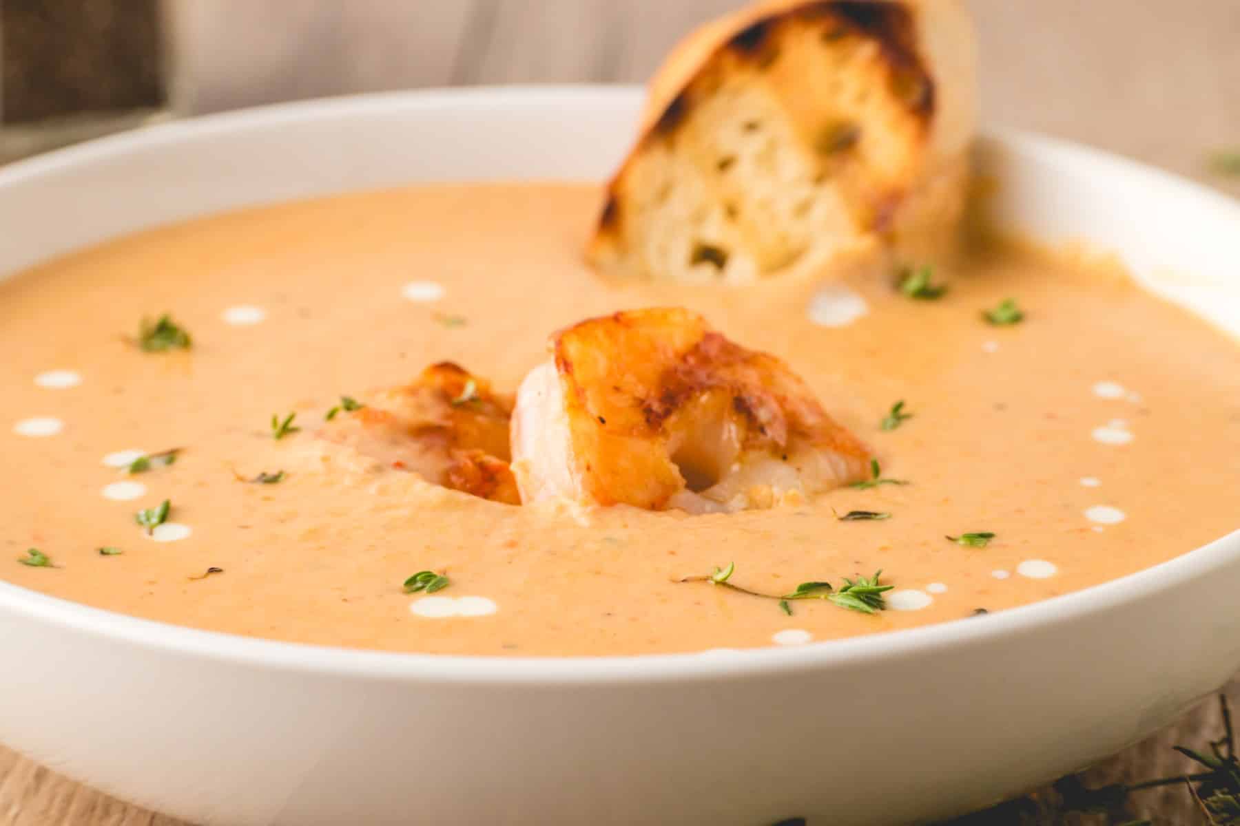 Horizontal picture of a bowl of shrimp bisque with shrimp on top and toasted bread dunking in. 