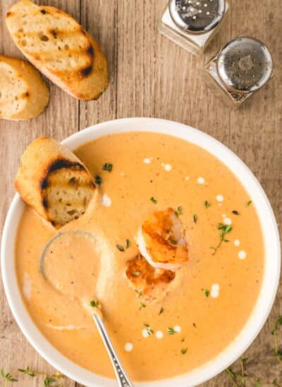 Overhead of a bowl of shrimp bisque with shrimp on top and dotted cream and a grilled piece of bread dunked in.