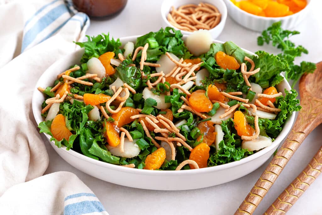 A colorful salad in a white oval dish featuring vibrant kale, mandarin orange slices, crunchy chow mein noodles, and water chestnuts. Wooden salad servers and a striped cloth are placed beside the dish, with bowls of additional toppings visible in the background.