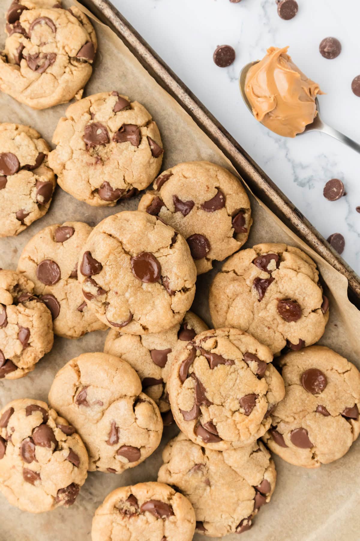 Peanut Butter Chocolate Chip Cookies on a baking sheet piled on top of each other with a spoonful of peanut butter and chocolate chips on the side. 