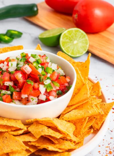 Bowl full of Pico de Gallo with tortilla chips and tomatoes, jalapenos and lime in the background.