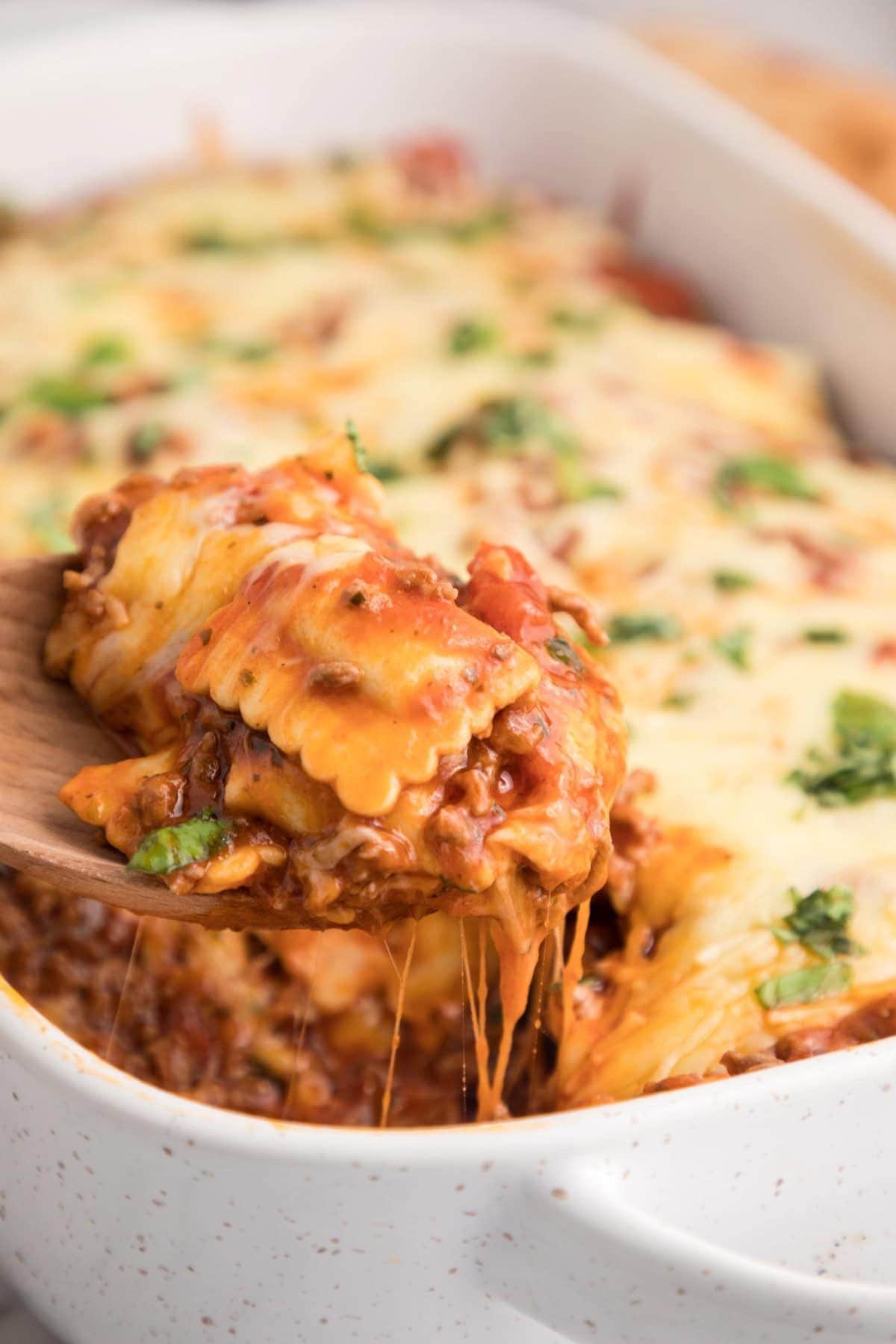 Wooden spoonful of baked ravioli casserole being removed from a casserole dish.