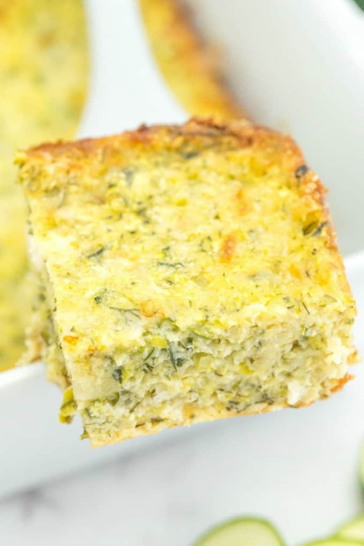 Slice of cheesy zucchini bake on a spatula held above a baking dish.