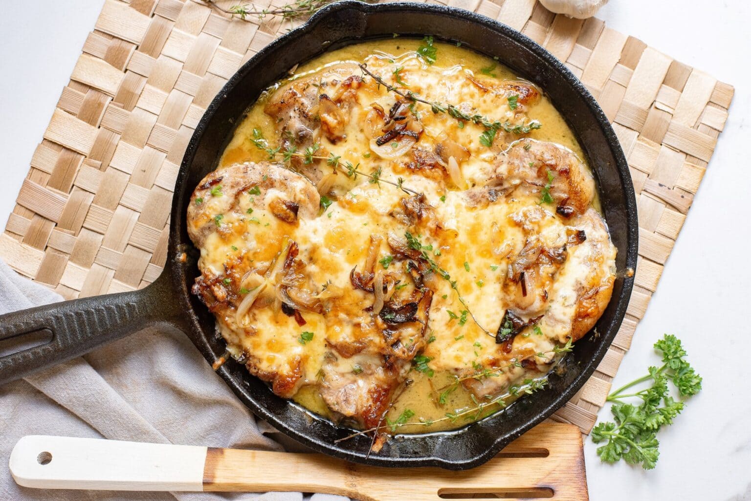 A cast iron skillet filled with cheesy, golden-baked pork chops topped with caramelized onions and fresh herbs.
