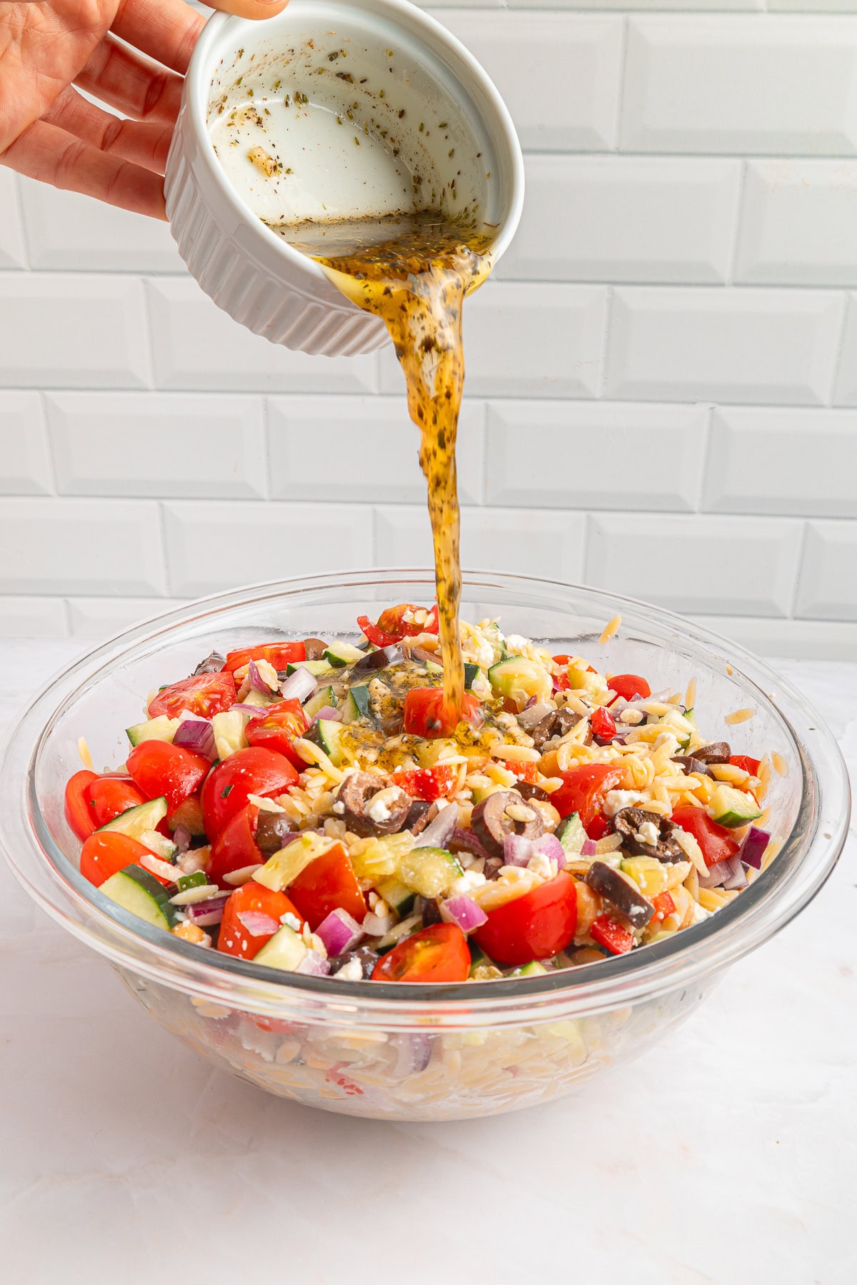 Another winner of a summer salad is this Greek Orzo Pasta Salad. It is easily portable, making it great for a picnic, BBQ or any gathering. It is hearty and healthy and full of flavors! Pouring dressing over the salad.